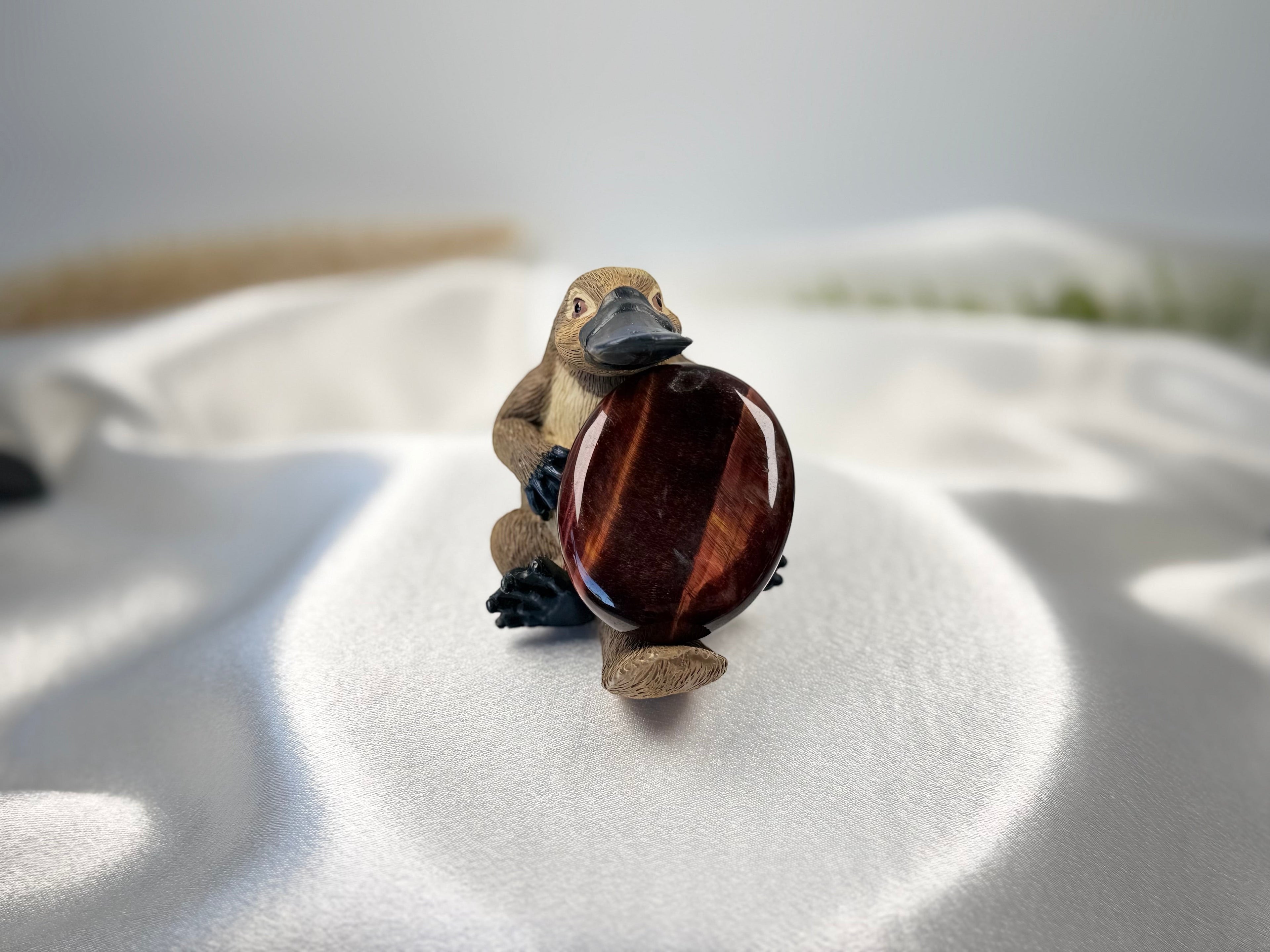 Treasured Energies red tiger's eye worry stone held by a small platypus figurine on a white satin background
