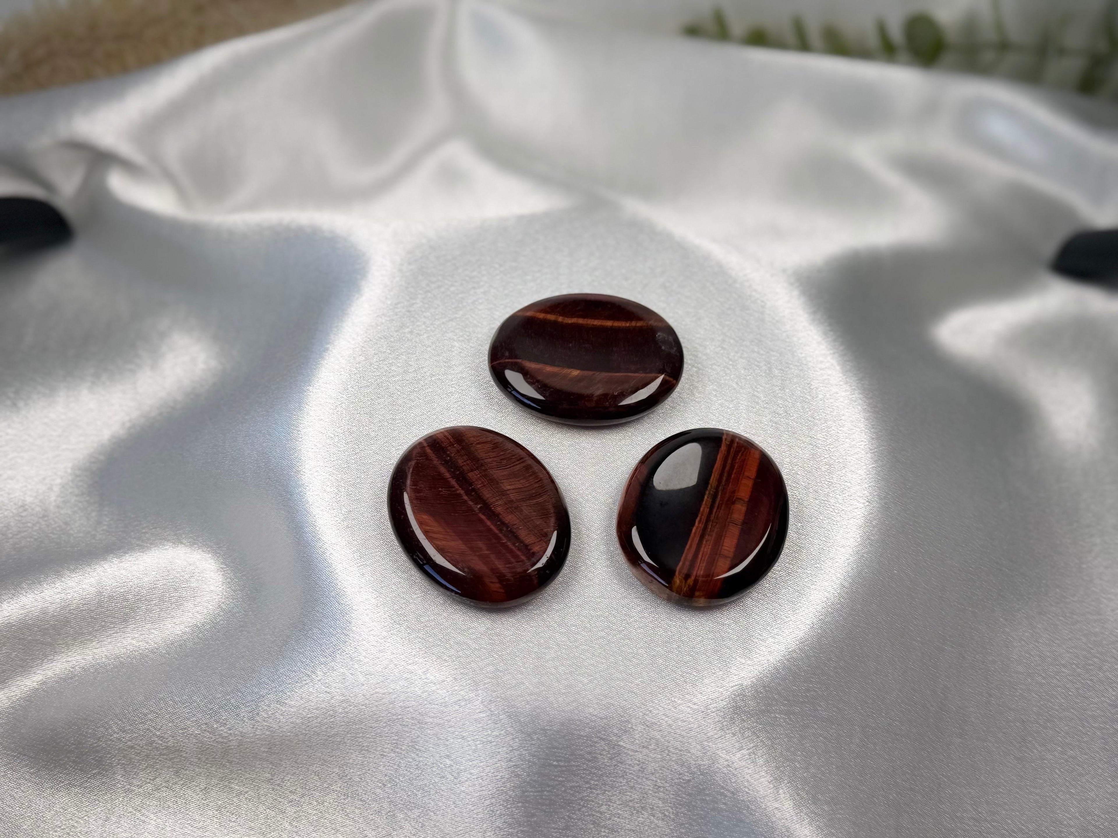 Treasured Energies red tiger's eye worry stones with smooth, polished surfaces resting on white satin fabric.