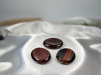 Three polished Red Tiger's Eye worry stones with deep red and brown banding on a soft white satin fabric background.