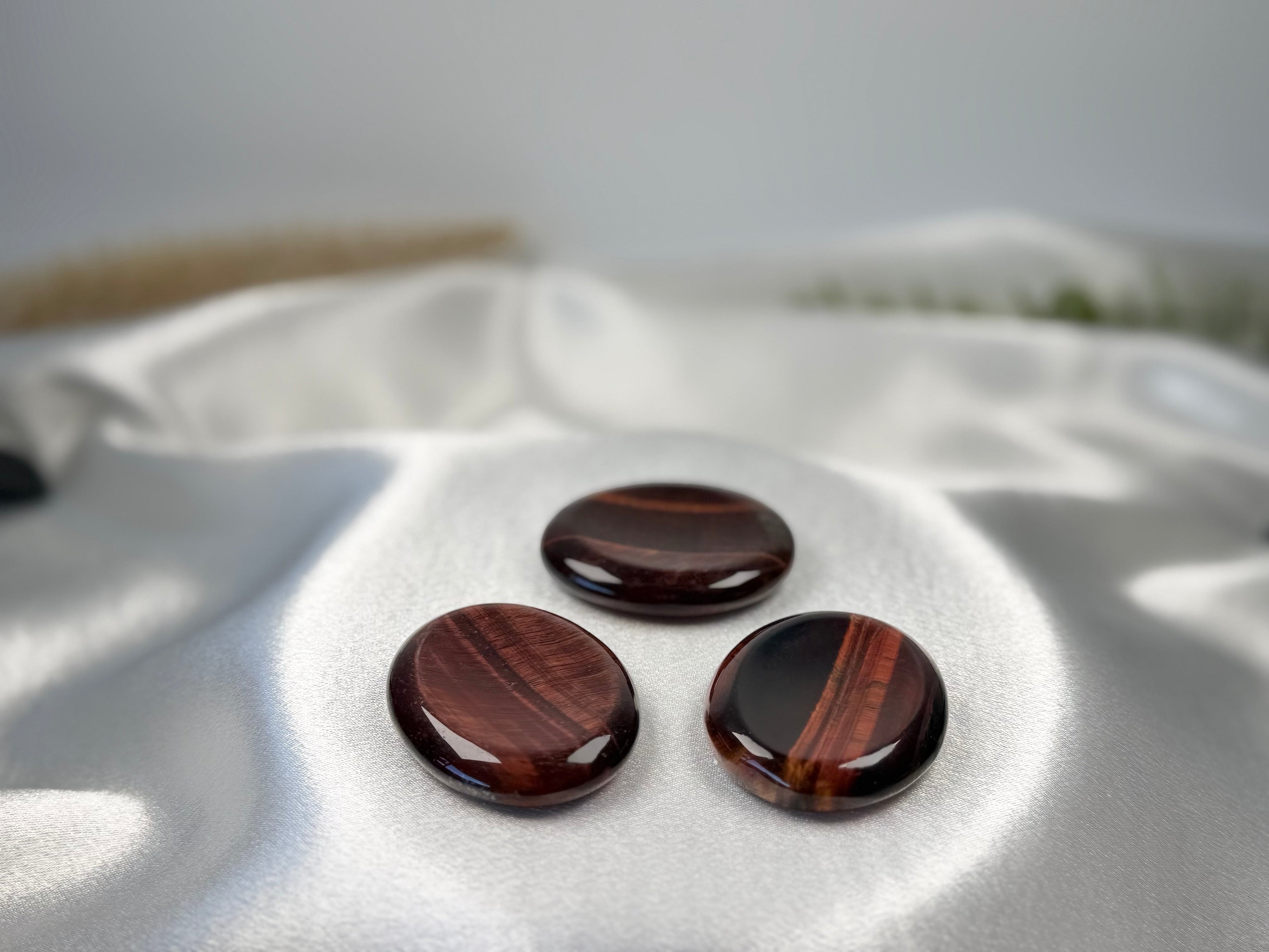 Three polished Red Tiger's Eye worry stones with deep red and brown banding on a soft white satin fabric background.