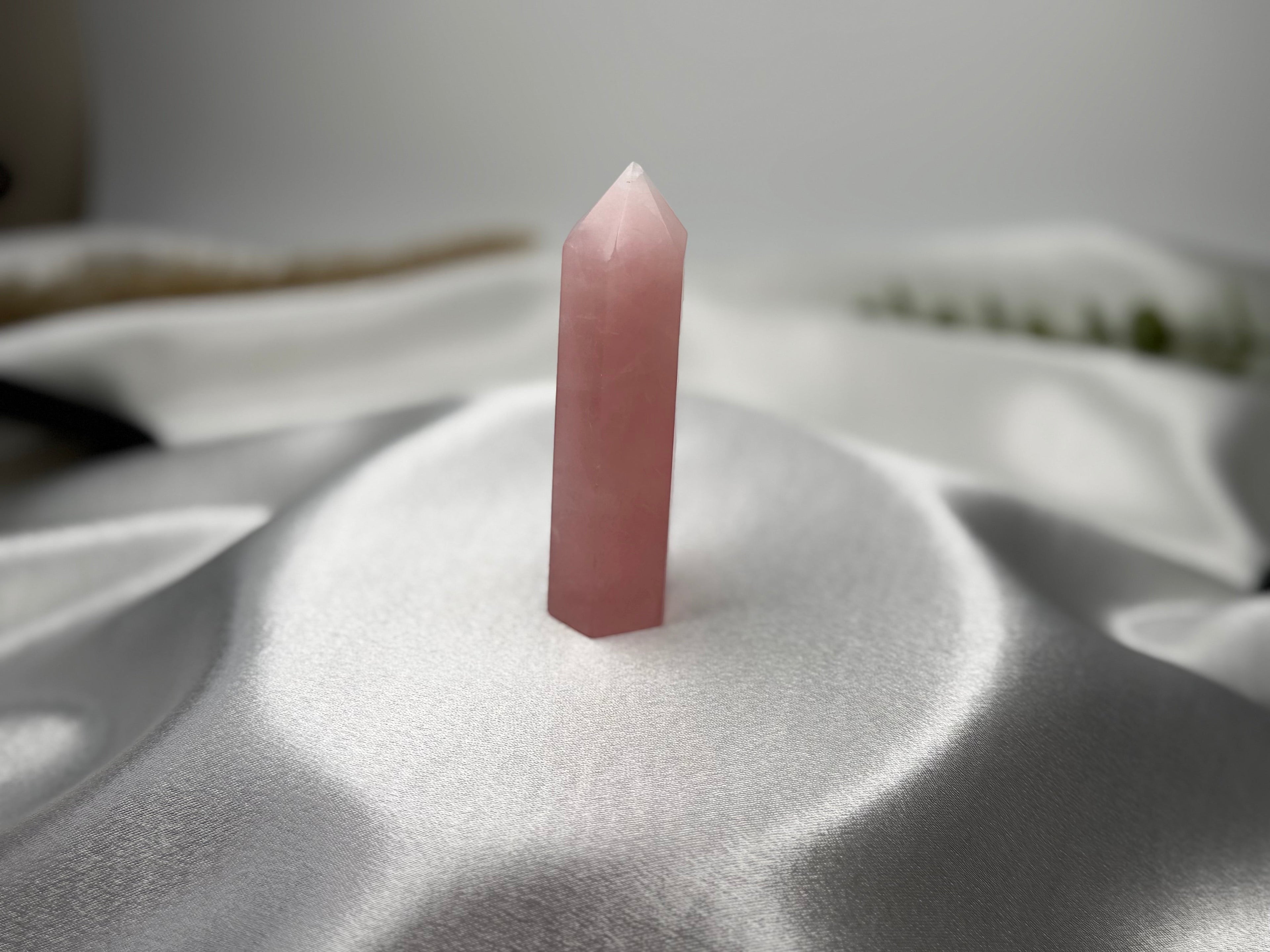 Treasured Energies rose quartz crystal tower with a pointed tip, displayed on a smooth white satin fabric background.