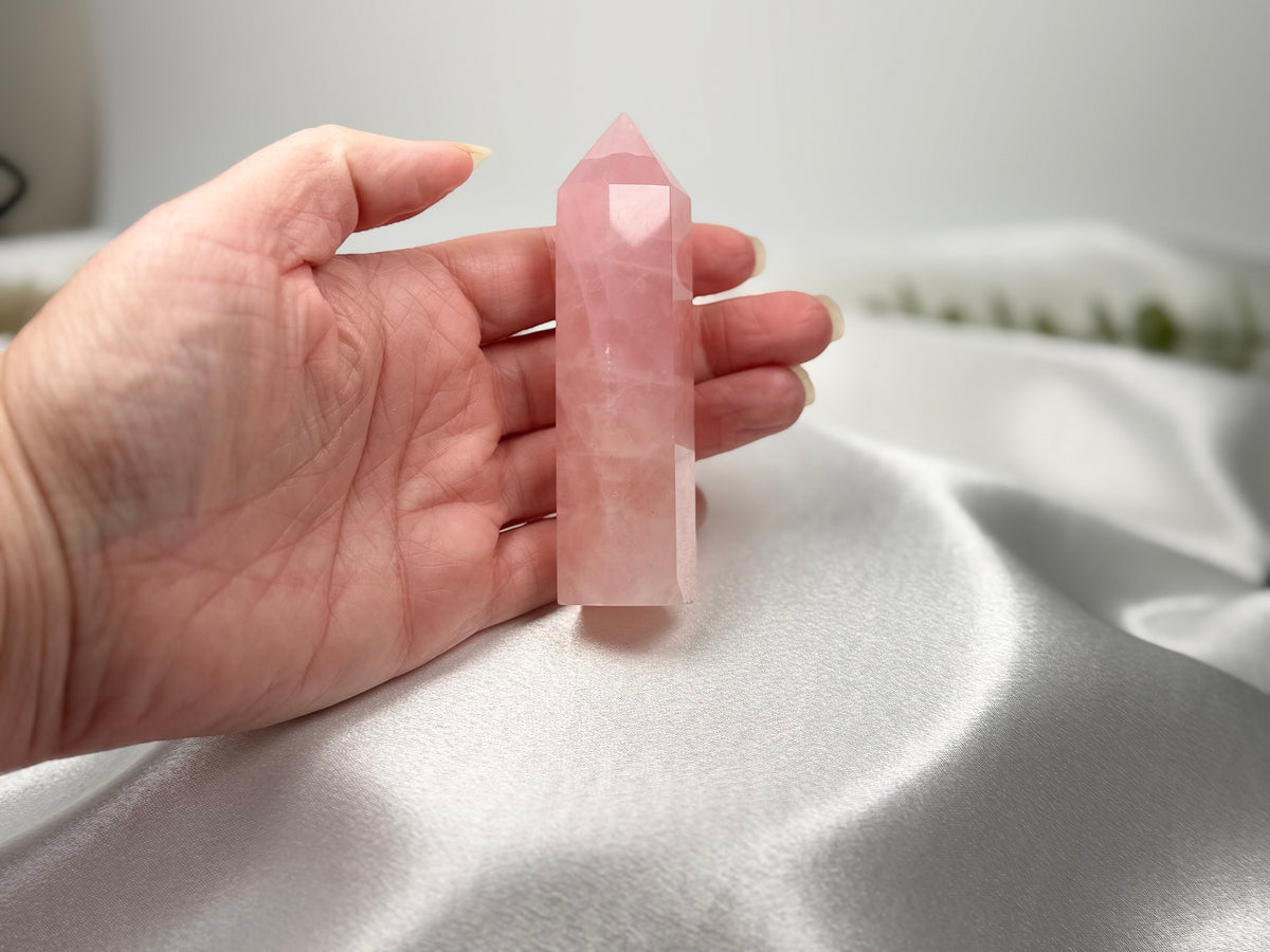 Treasured Energies polished rose quartz crystal generator tower held in a hand against a soft white satin background.