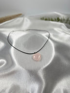 Treasured Energies heart-shaped rose quartz crystal pendant on a simple black cord necklace against white satin.