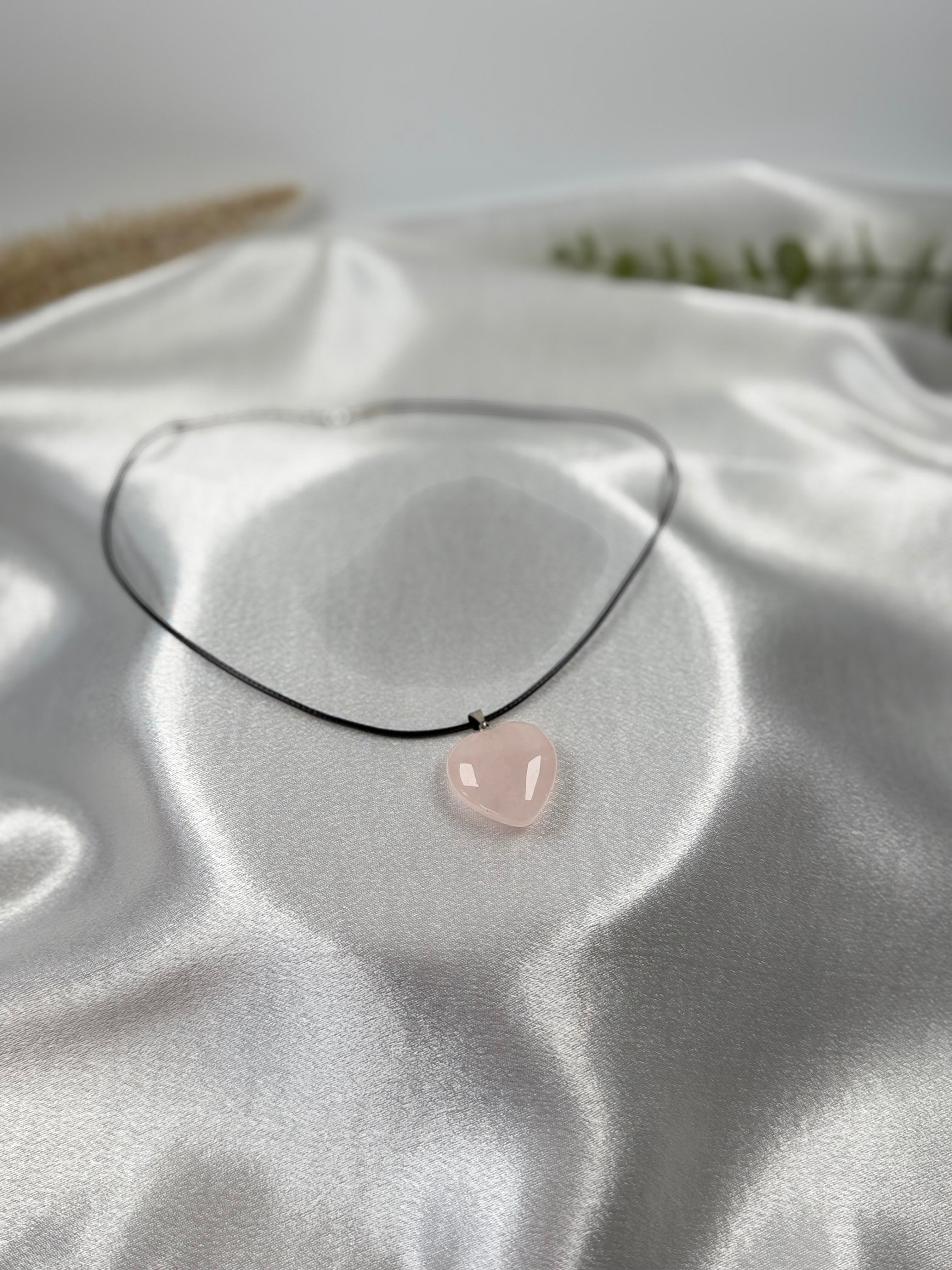 Treasured Energies heart-shaped rose quartz crystal pendant on a simple black cord necklace against white satin.
