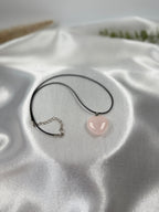 Treasured Energies heart-shaped rose quartz pendant on a black cord necklace resting on white satin fabric.
