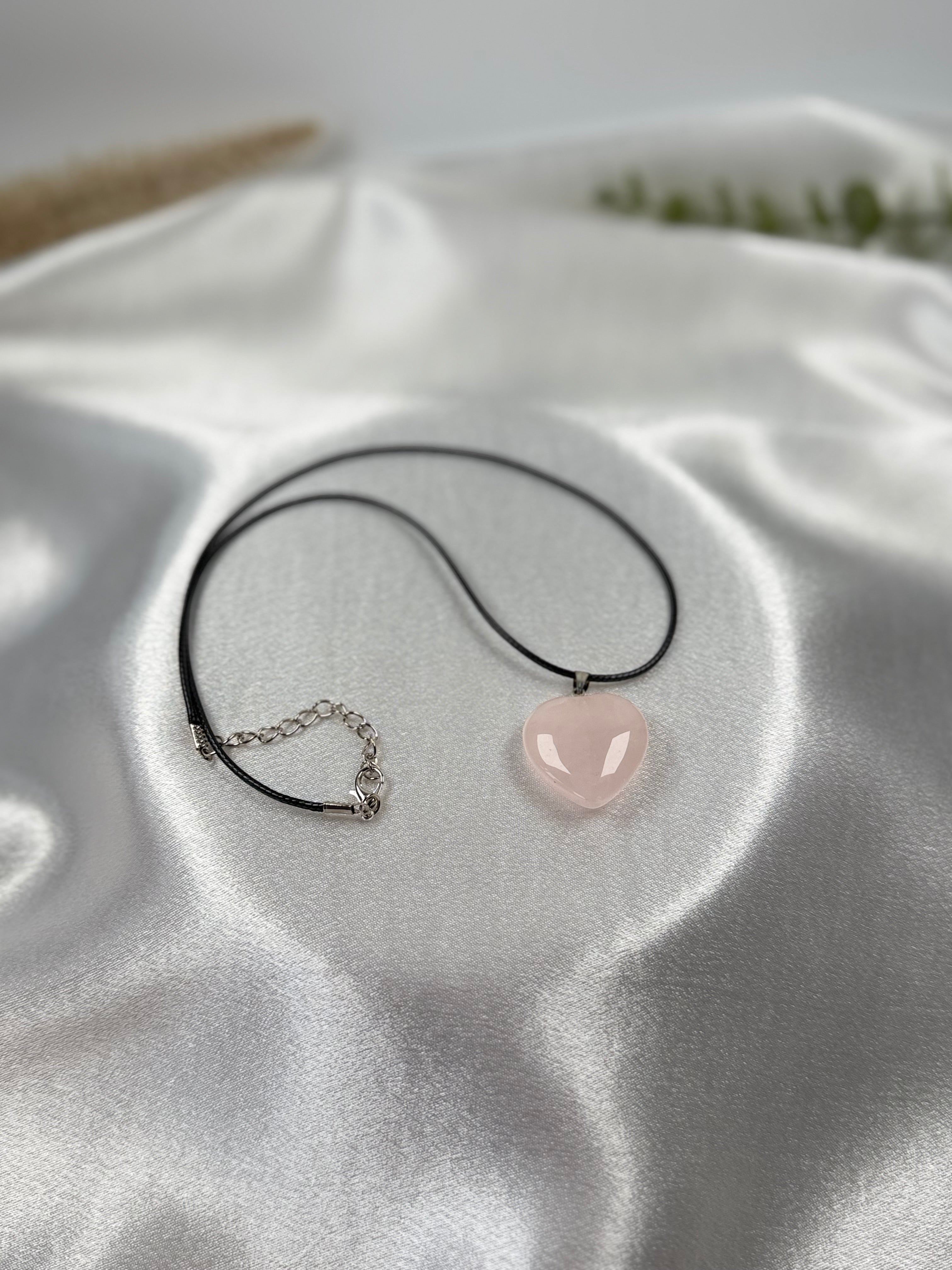 Treasured Energies heart-shaped rose quartz pendant on a black cord necklace resting on white satin fabric.