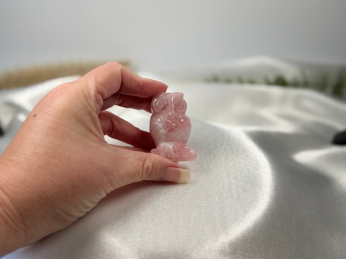 Treasured Energies rose quartz owl carving held in a hand against a soft white satin background.