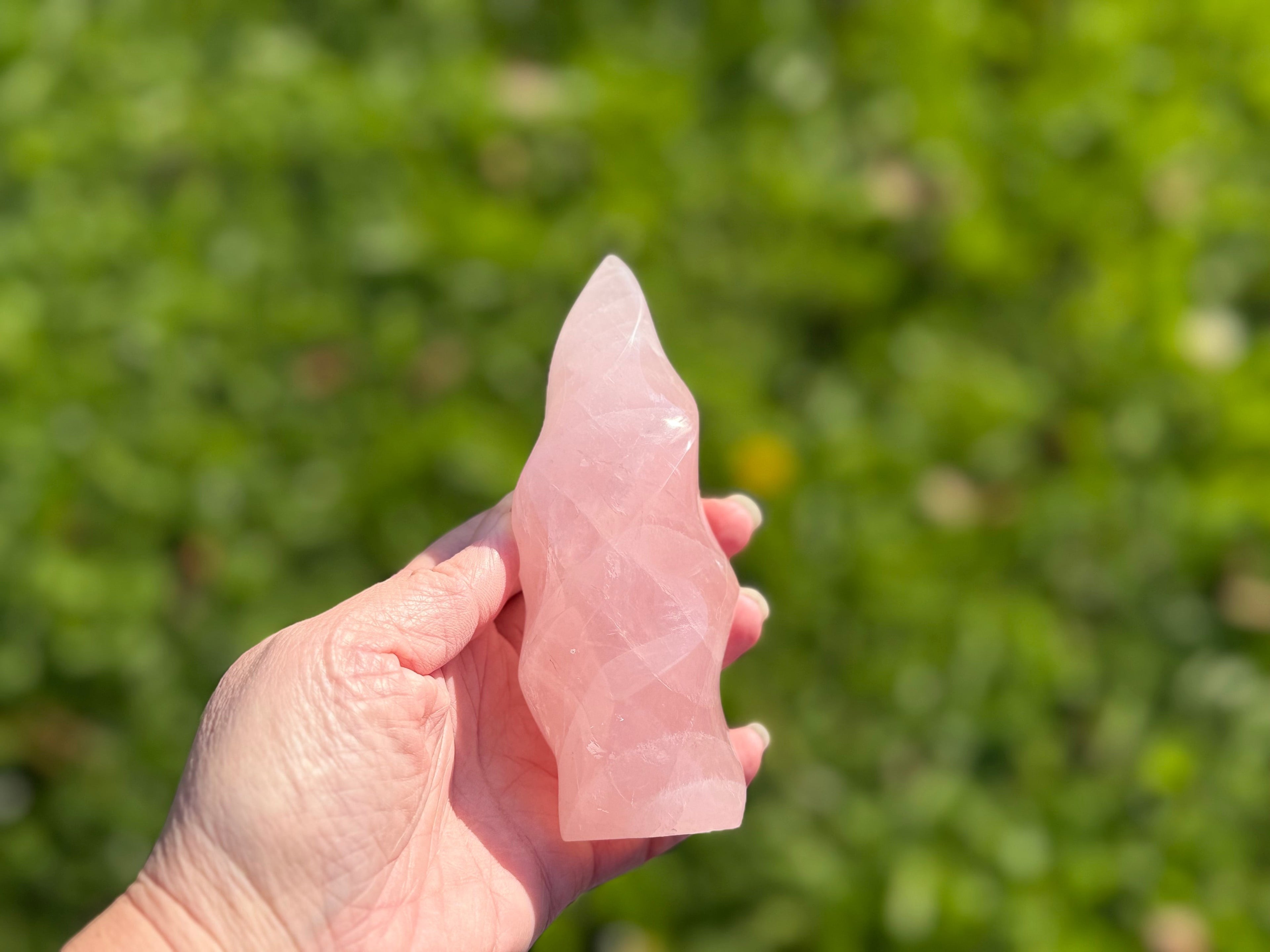 Treasured Energies large rose quartz crystal carved into a smooth, spiraling flame shape held in a hand outdoors.
