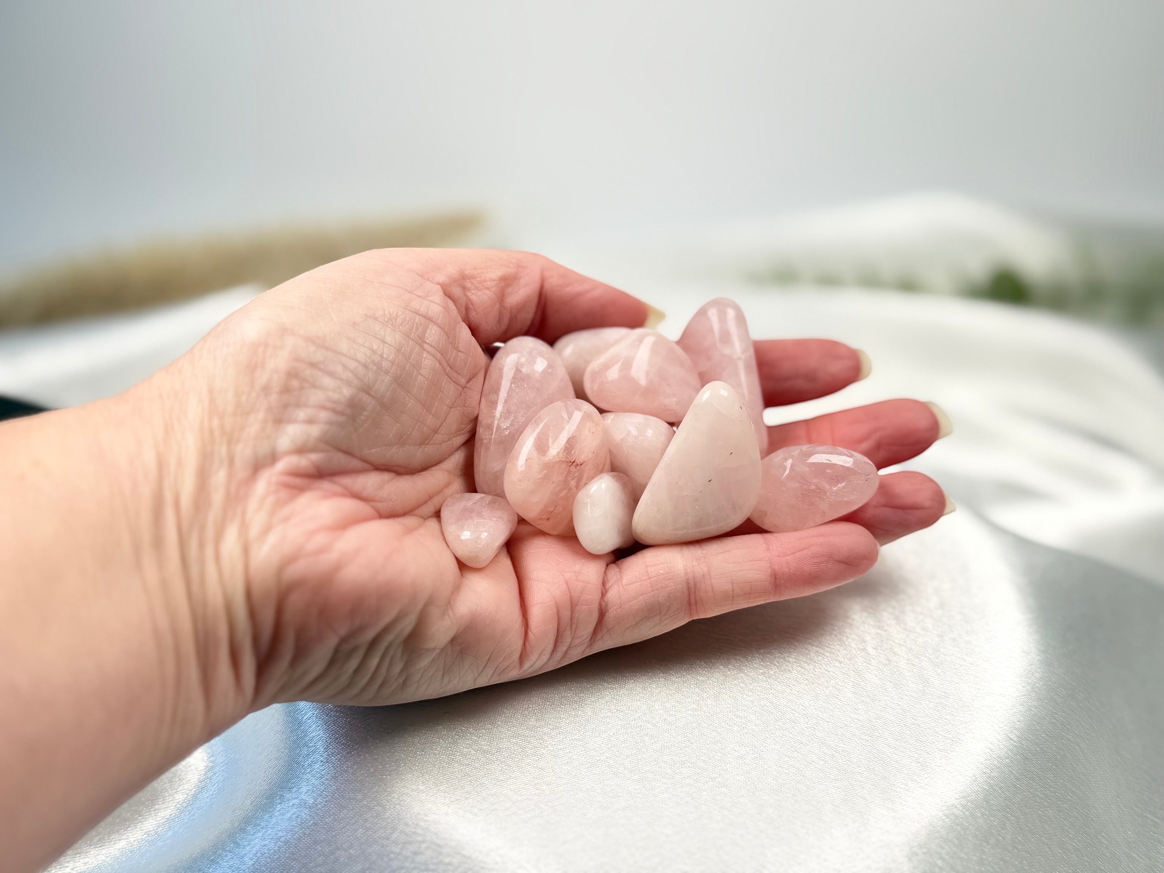 Treasured Energies hand holding several smooth, polished pale pink rose quartz tumbled crystals on white silk fabric.