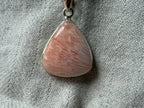 Treasured Energies Scolecite crystal pendant in a polished sterling silver setting on a textured grey fabric background.