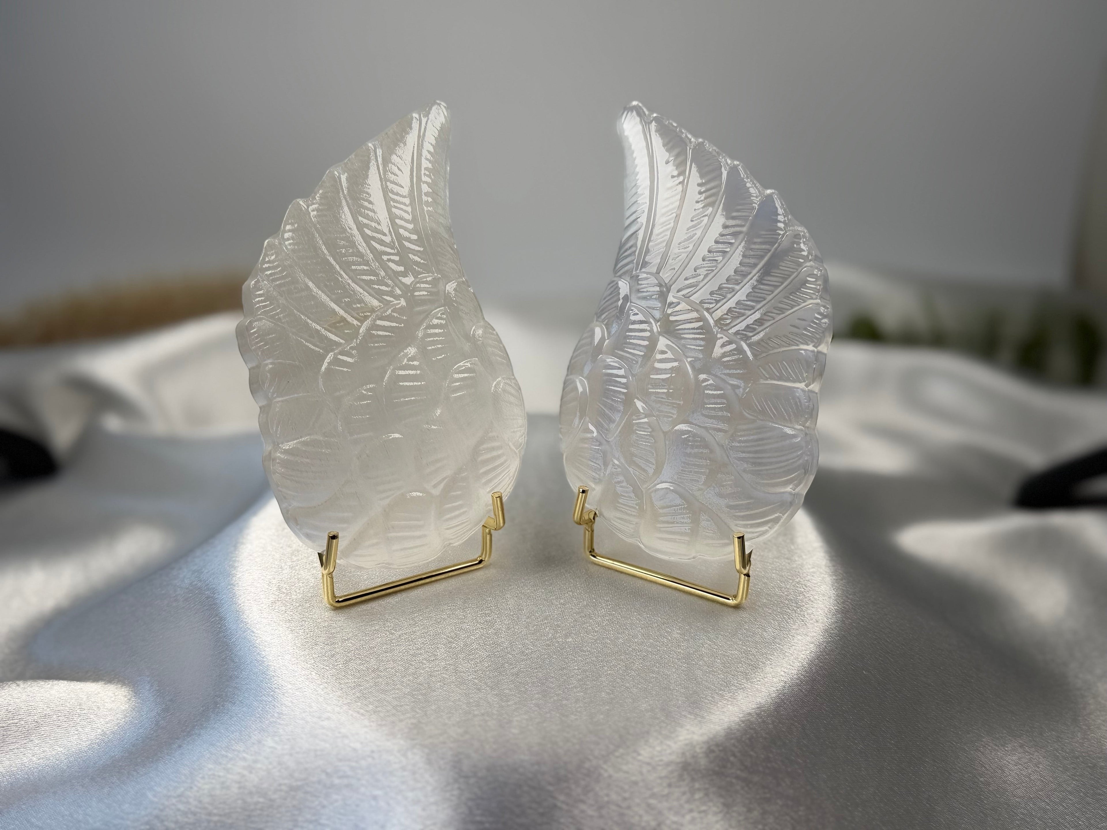 Treasured Energies pair of carved white selenite angel wings on gold stands displayed on a satin fabric surface.