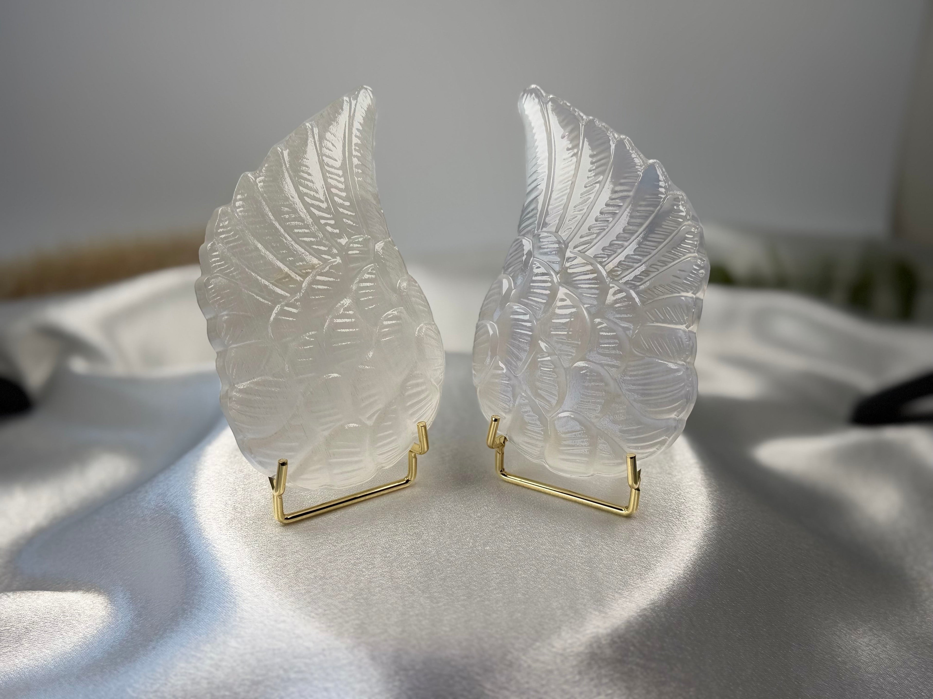 Treasured Energies pair of carved white selenite angel wings on small gold stands against a white satin background.