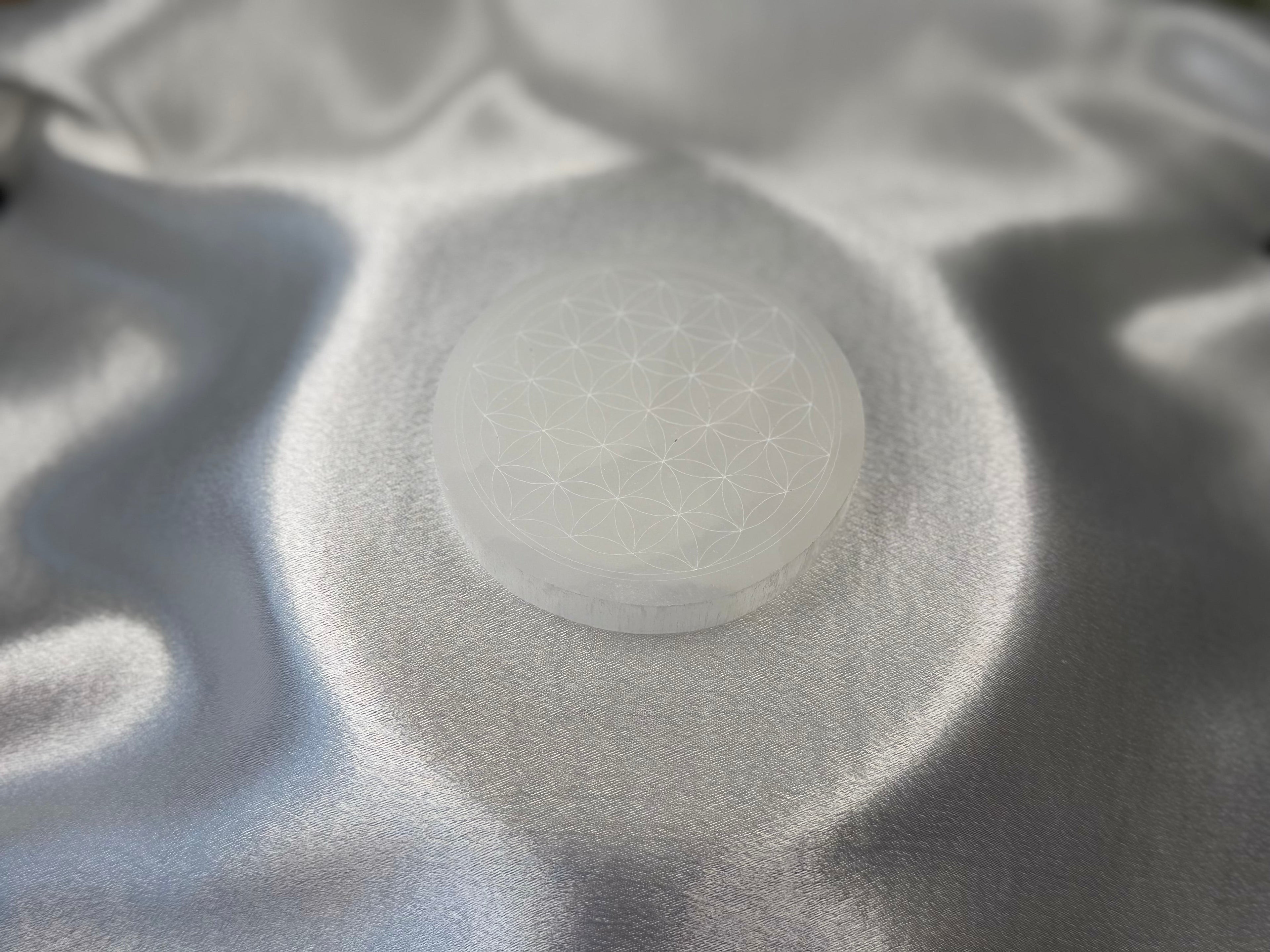 Treasured Energies 8cm round white selenite charging plate with a delicate engraved flower of life pattern on satin