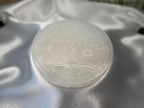 Treasured Energies 14cm round white selenite charging plate with an engraved tree of life design on satin fabric.