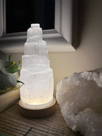 Treasured Energies tiered selenite crystal tower lamp glowing with soft light on a wooden base next to a geode