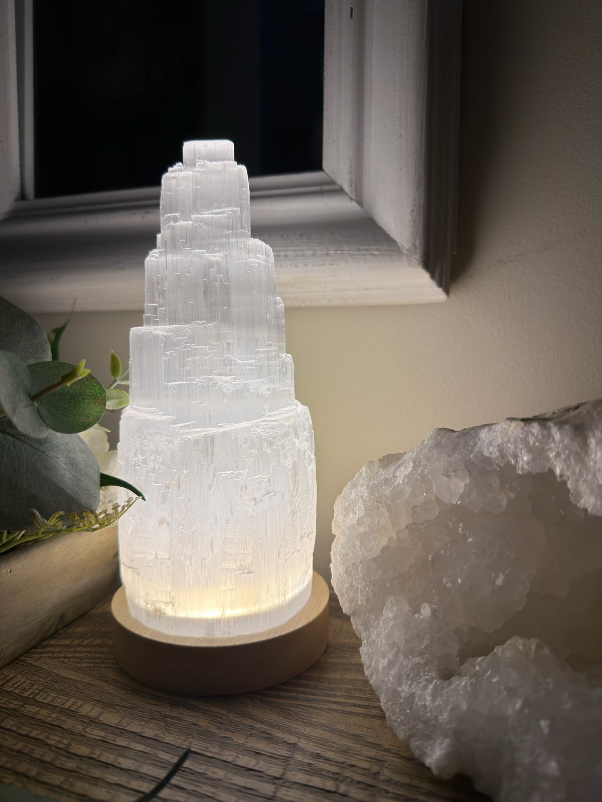 Treasured Energies tiered selenite crystal tower lamp glowing with soft light on a wooden base next to a geode