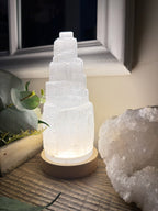 Treasured Energies selenite crystal tower lamp with a tiered design and warm LED glow on a natural timber base.