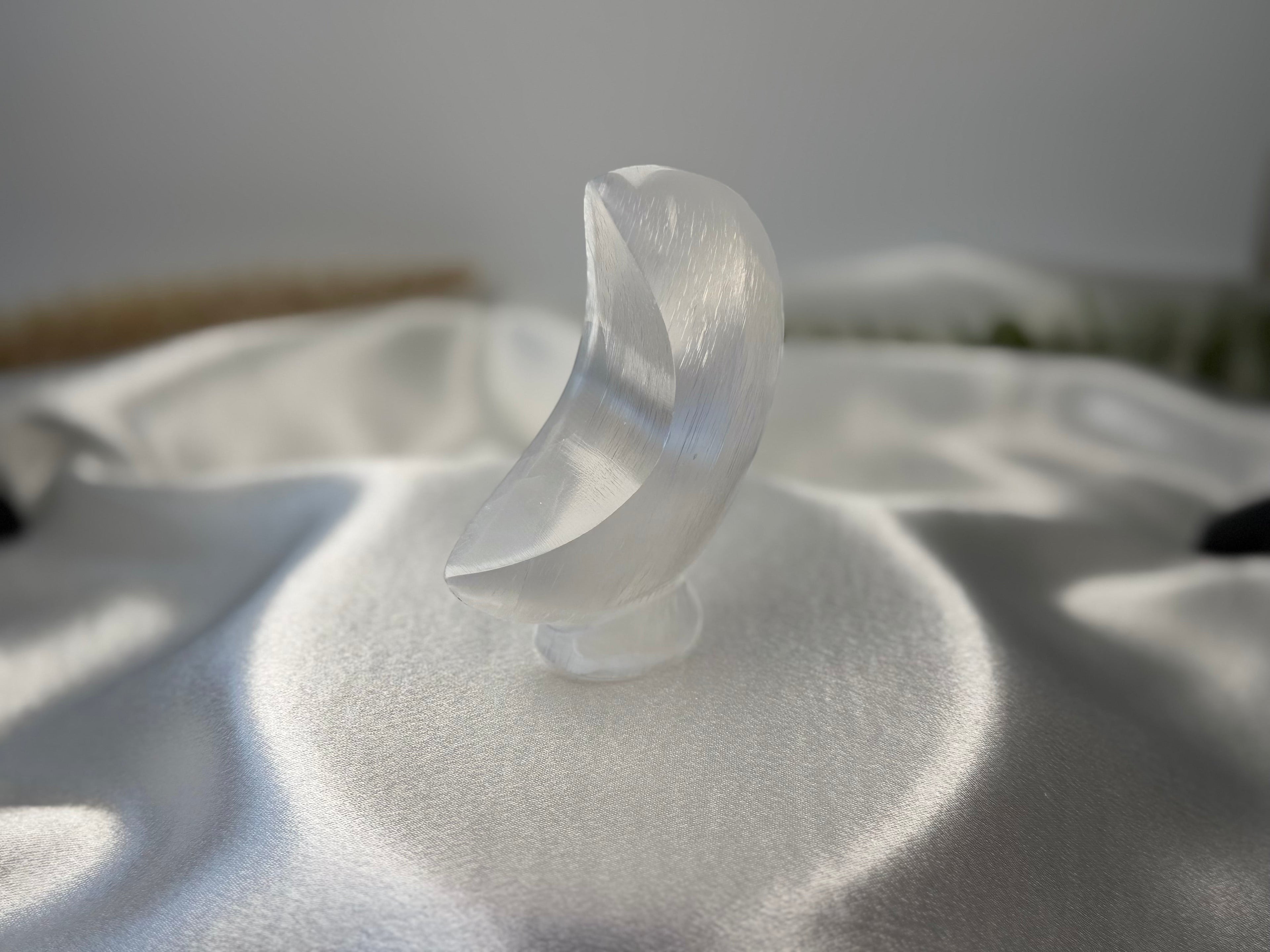 Treasured Energies hand-carved white selenite crescent moon crystal sculpture resting on a white satin fabric surface.