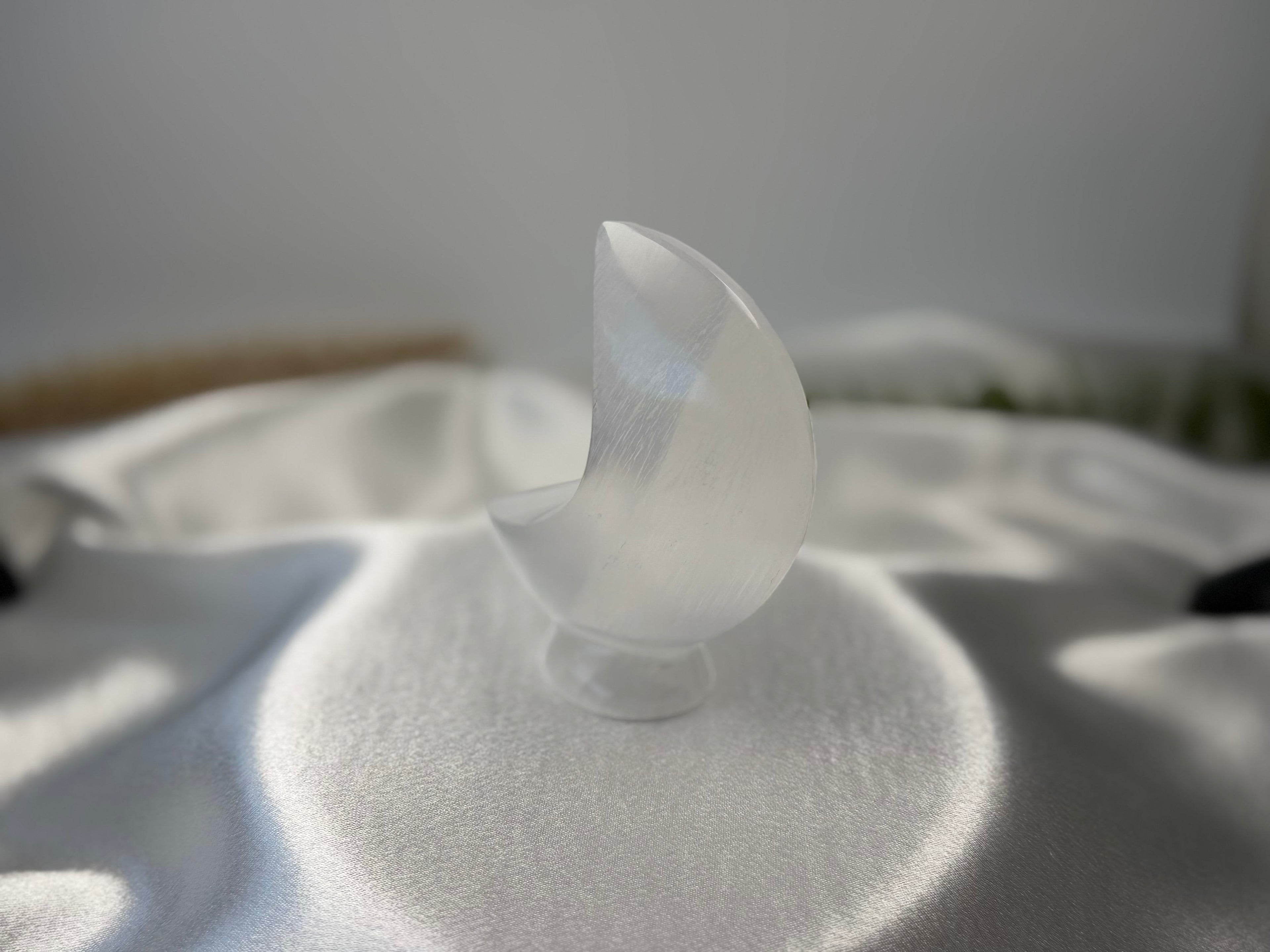 Treasured Energies hand-carved white selenite crescent moon crystal sculpture resting on a soft white satin fabric.
