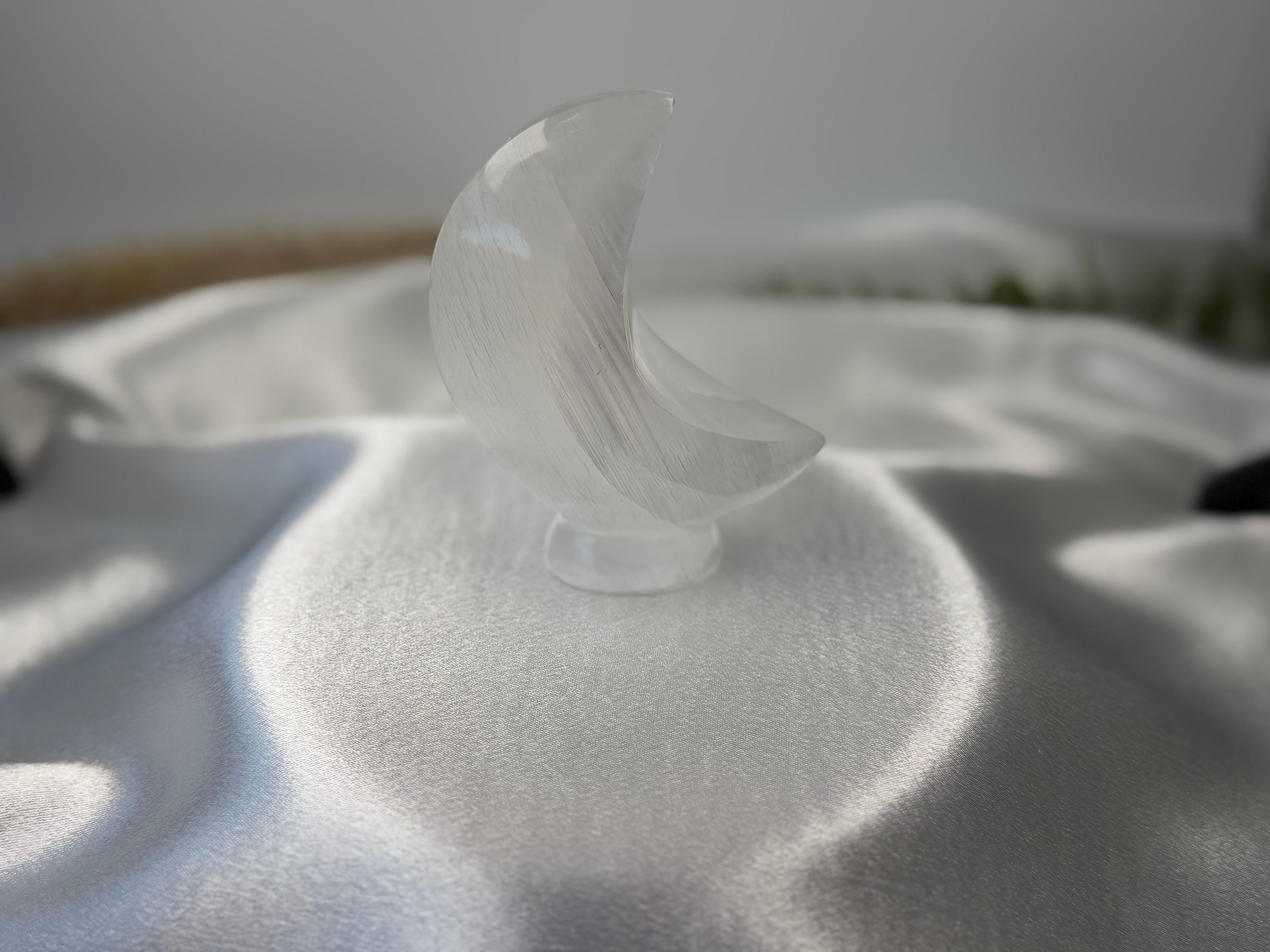 Treasured Energies hand-carved translucent white selenite crescent moon crystal sculpture on a satin fabric background.
