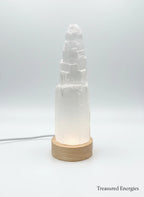 Treasured Energies small selenite crystal tower lamp with a round wooden LED base glowing against a white background.