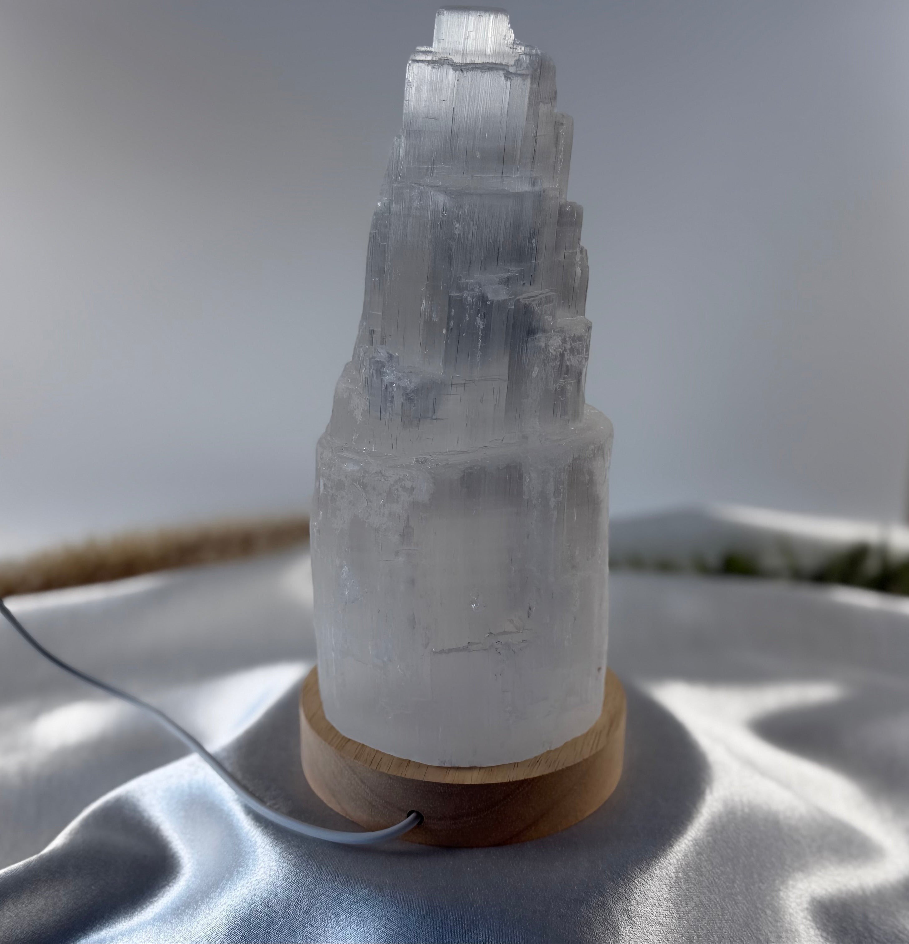 Treasured Energies small selenite crystal tower lamp with a natural wood LED base on a white satin fabric background.