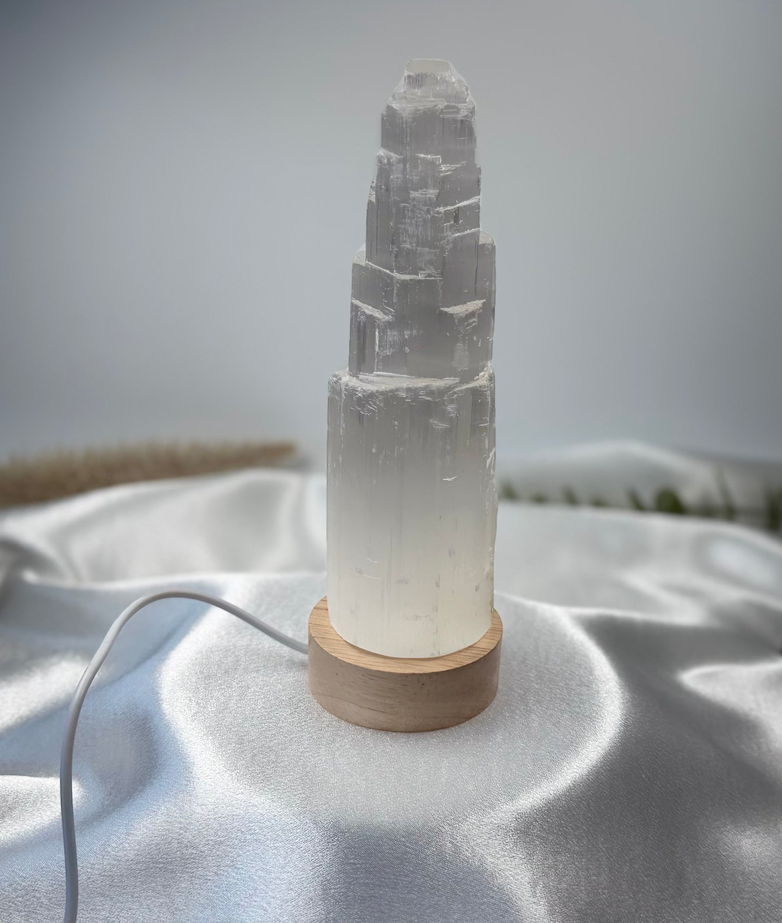 Treasured Energies small selenite tower lamp with a natural wood LED base on a white satin fabric background.