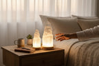 Treasured Energies small selenite tower lamp with LED base glowing on a wooden nightstand in a cozy bedroom setting
