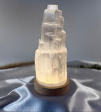 Treasured Energies small selenite tower lamp with a soft glowing LED light on a wooden base against a satin background.