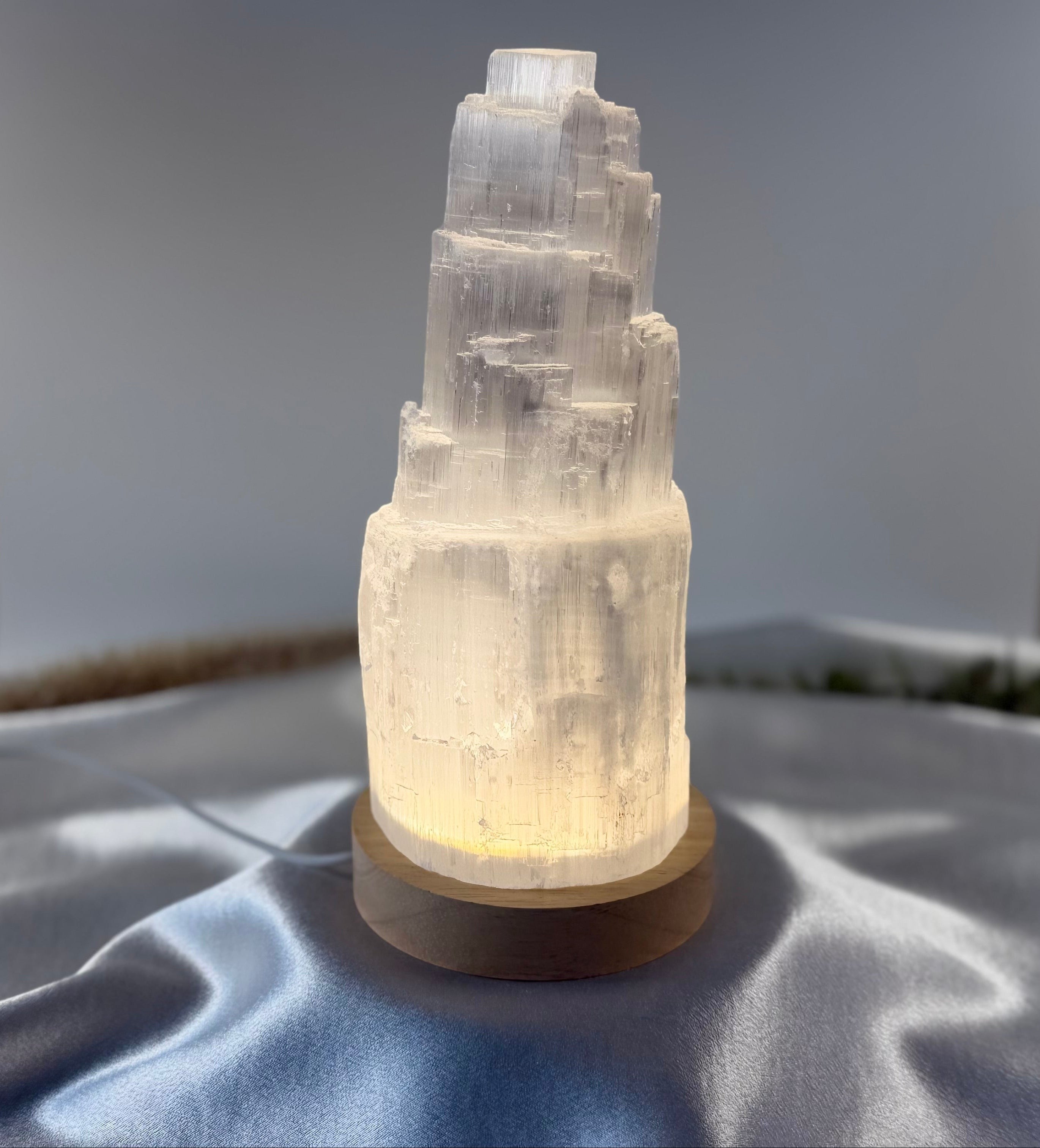 Treasured Energies small selenite tower lamp with a soft glowing LED light on a wooden base against a satin background.