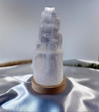 Treasured Energies small selenite crystal tower lamp on a round wooden LED base against a soft white fabric background.