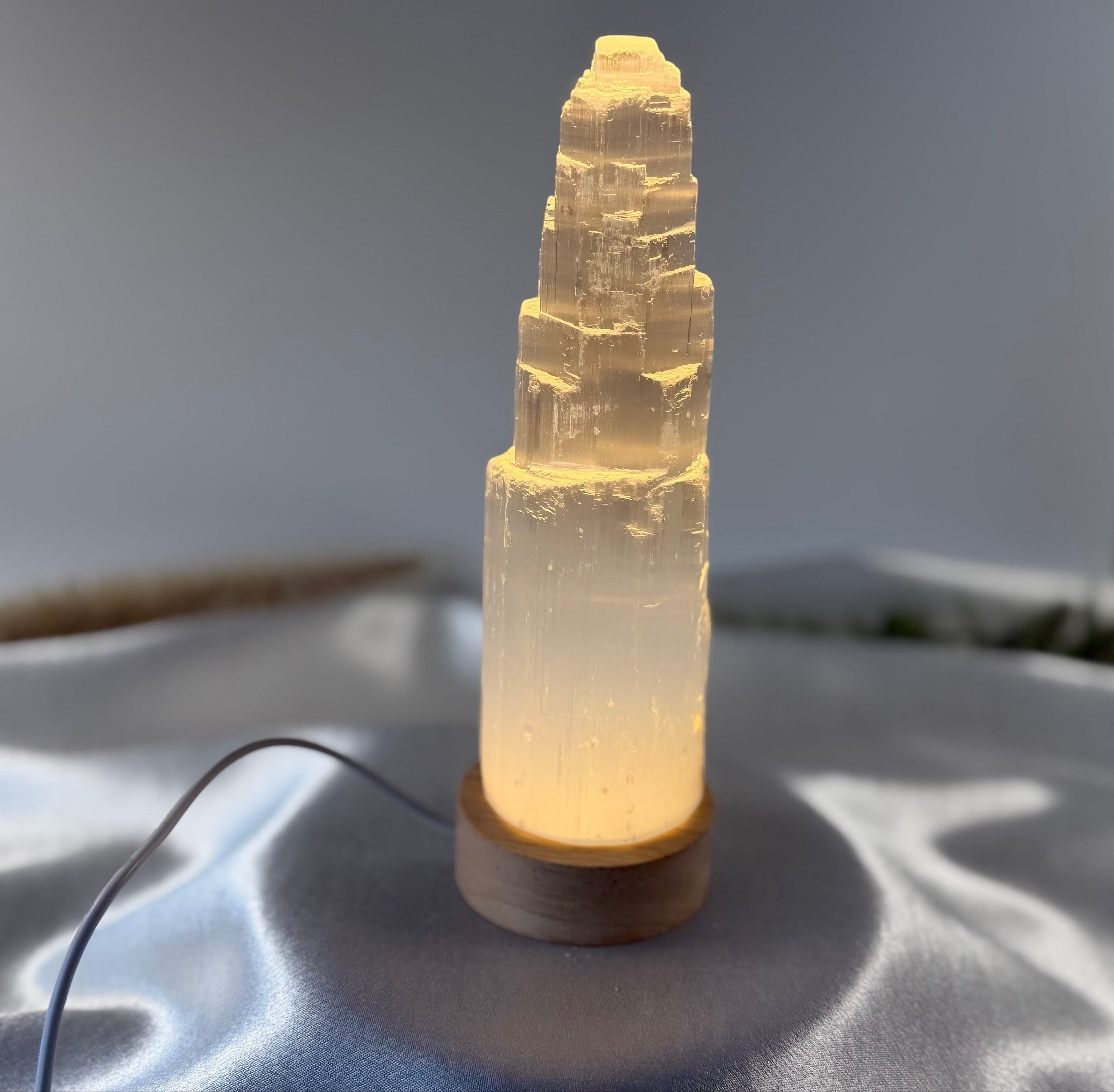 Treasured Energies small selenite tower lamp with a warm LED light base on a soft grey fabric background.