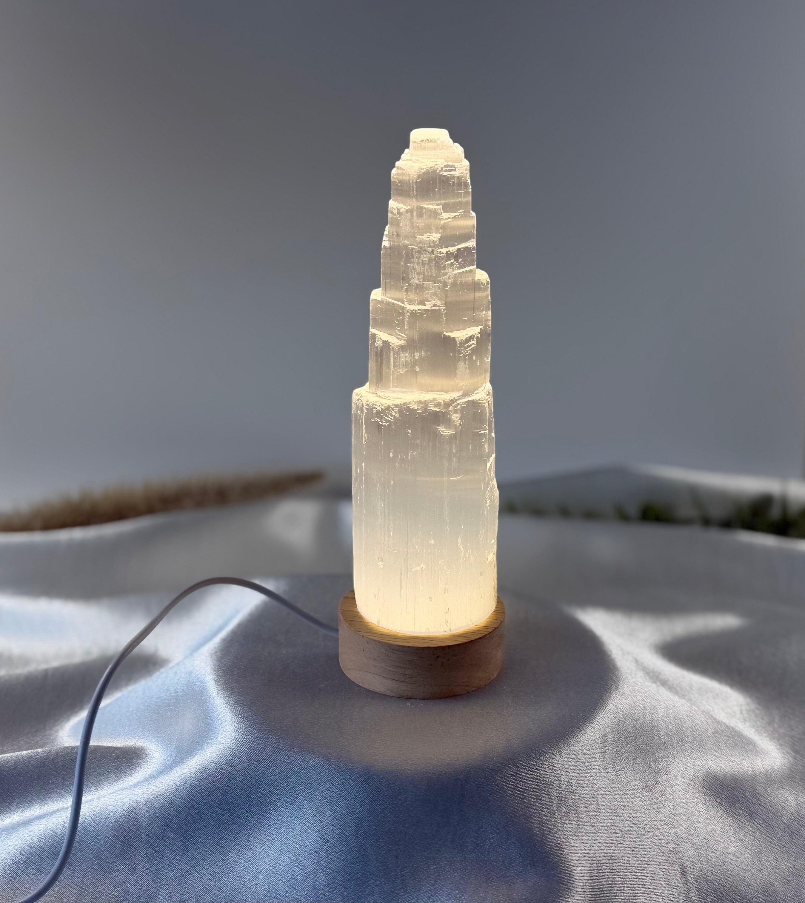 Treasured Energies small selenite tower lamp with a soft glowing LED wooden base on a draped satin background.