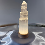 Treasured Energies small selenite crystal tower lamp with a glowing LED wooden base on a soft grey fabric surface.