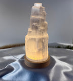 Treasured Energies small selenite tower lamp with a warm LED light on a wooden base, set on a soft grey fabric.