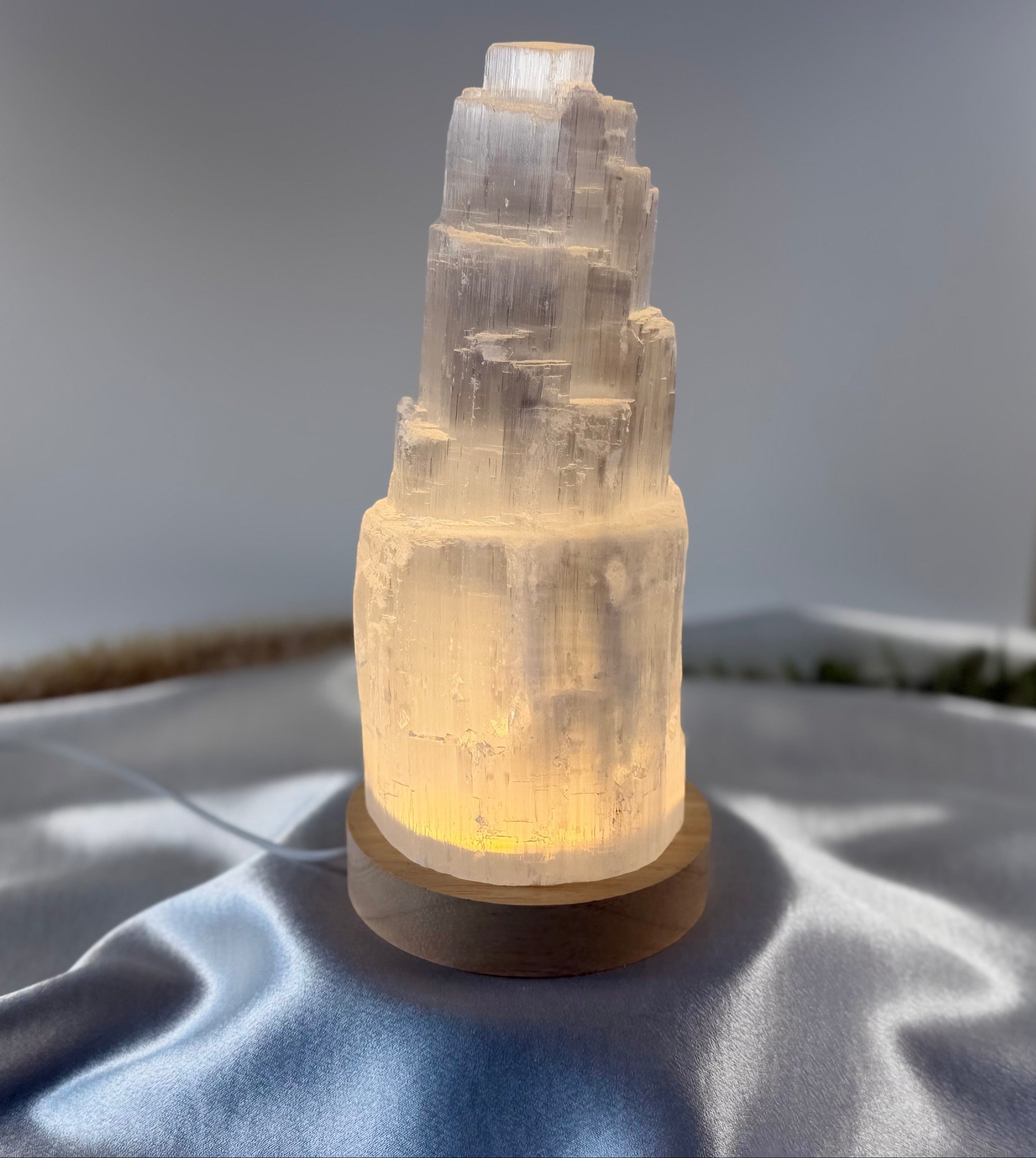 Treasured Energies small selenite tower lamp with a warm LED light on a wooden base, set on a soft grey fabric.