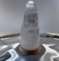 Treasured Energies small selenite tower lamp with a natural wood LED base on a white satin fabric background.