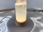 Treasured Energies small selenite tower lamp with a warm glowing LED base on a soft white fabric surface
