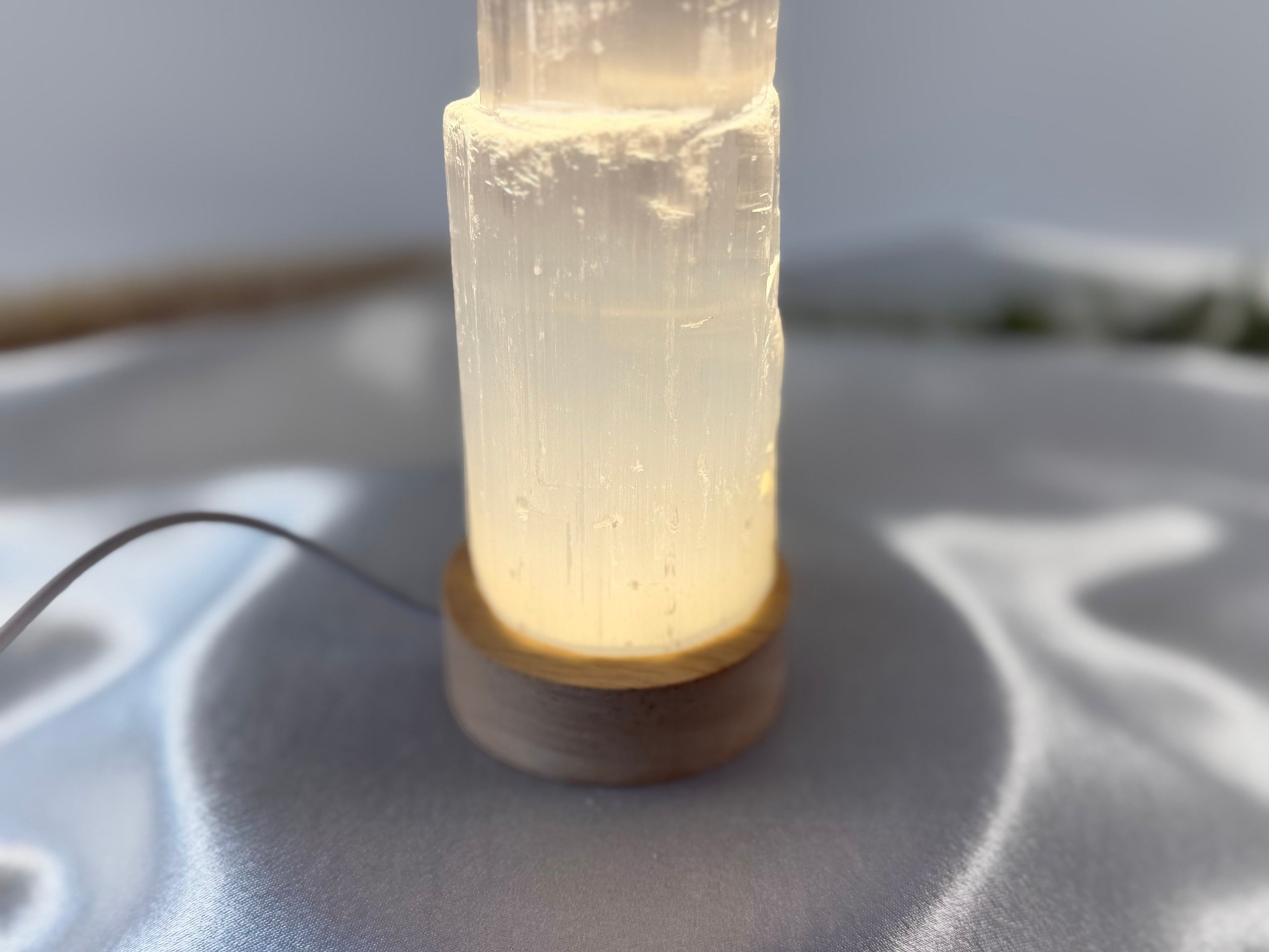 Treasured Energies small selenite tower lamp with a warm glowing LED base on a soft white fabric surface