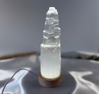 Treasured Energies small selenite crystal tower lamp with a round wooden LED base glowing on a soft fabric surface.