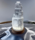 Treasured Energies small selenite tower lamp with a glowing LED light on a round wooden base against satin fabric.