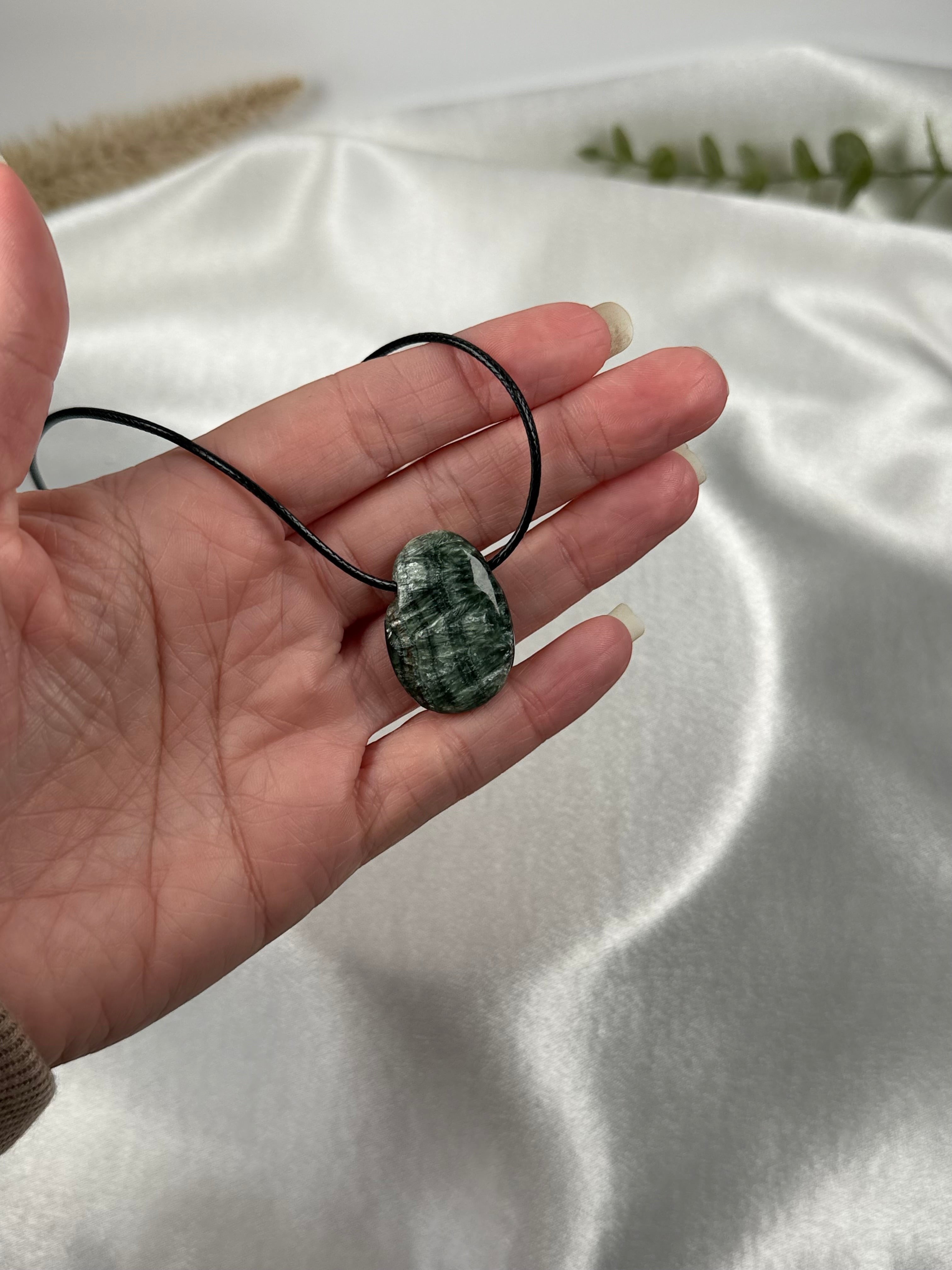 Treasured Energies seraphinite crystal pendant on a black cord necklace held in a hand against a white satin cloth.