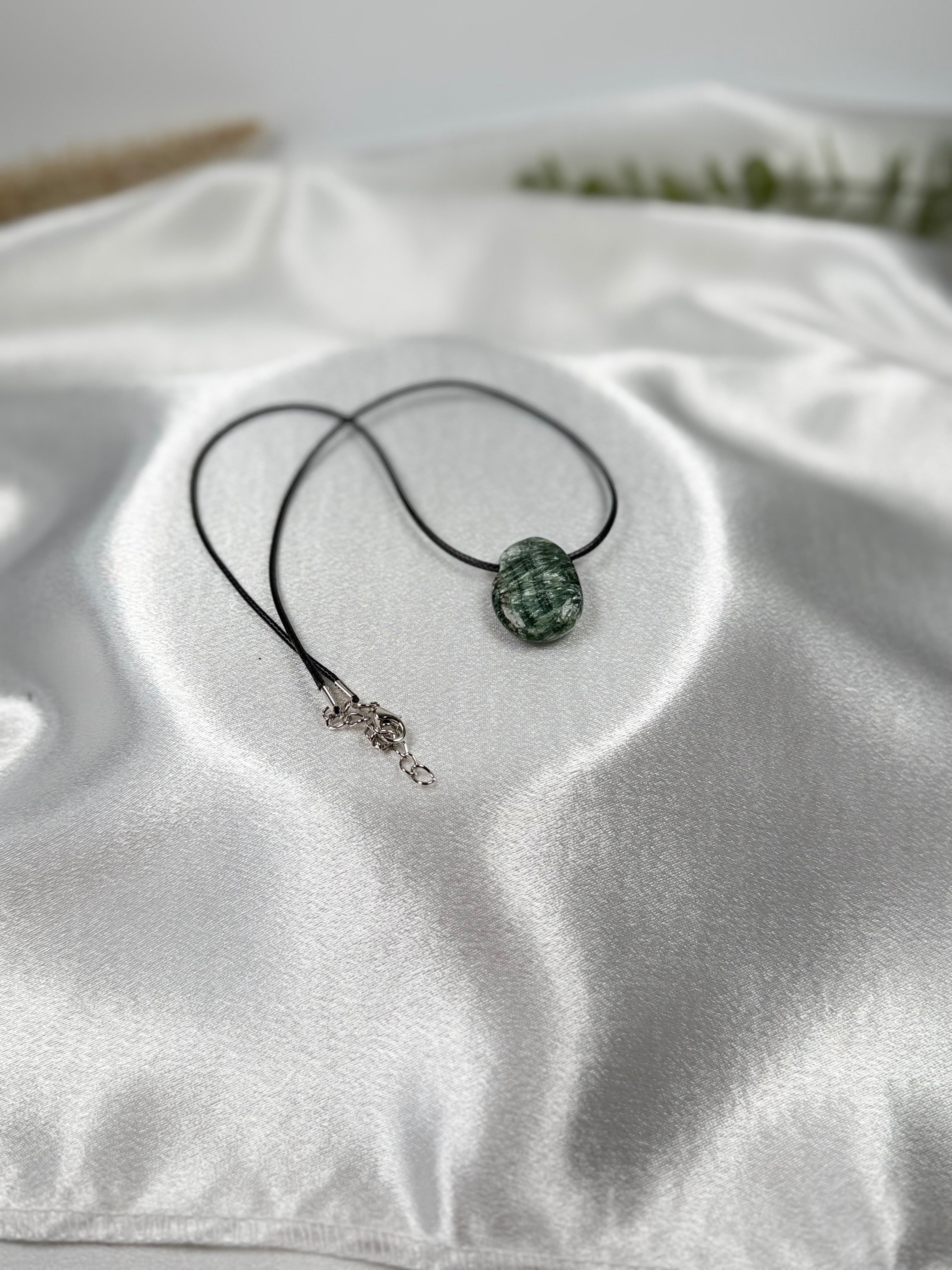 Treasured Energies seraphinite crystal pendant on a black cord necklace resting on white satin fabric