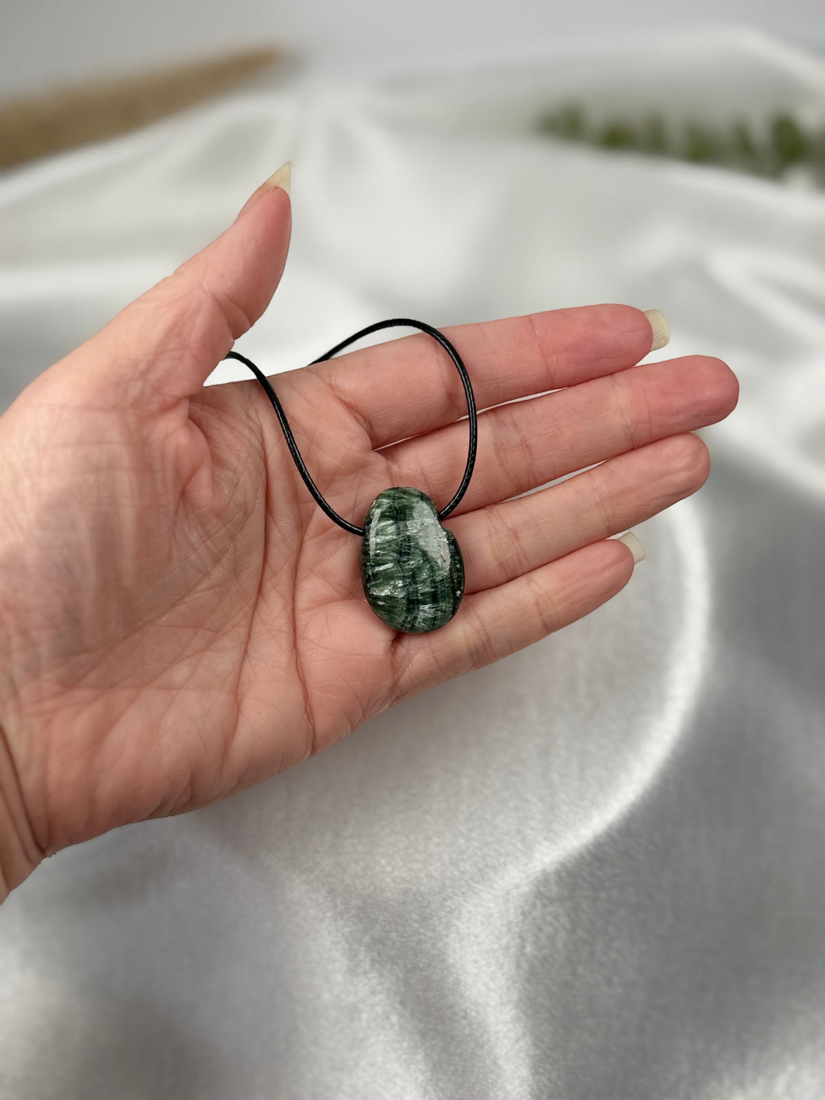 Treasured Energies seraphinite crystal pendant on a black cord held in a palm against a white silk background