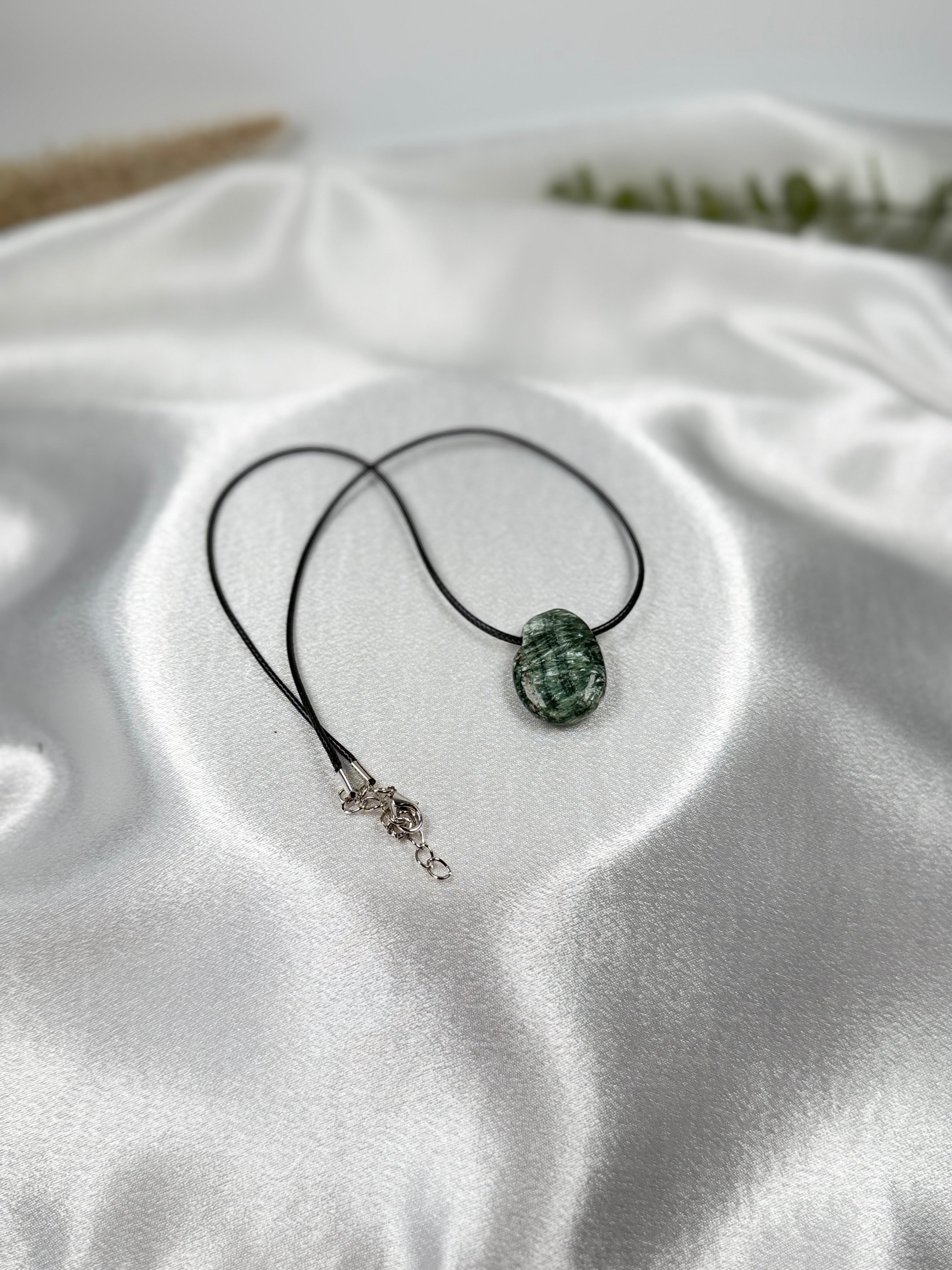 Treasured Energies seraphinite crystal pendant on a simple black cord necklace resting on white satin fabric.