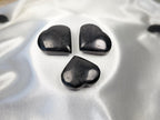 Three polished black shungite heart stones arranged on a smooth white satin fabric by Treasured Energies.