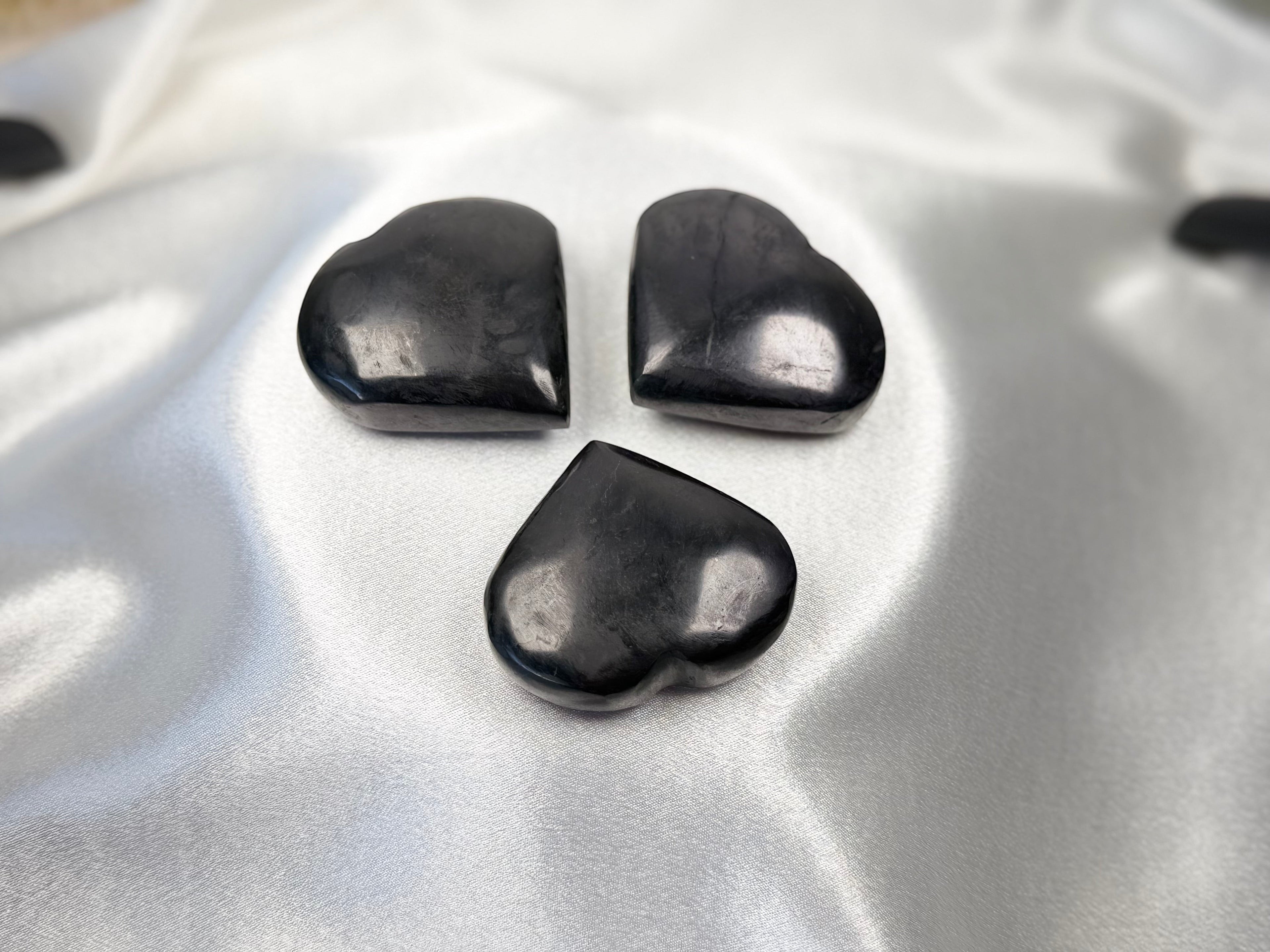 Three polished black shungite heart stones arranged on a smooth white satin fabric by Treasured Energies.