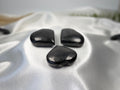 Three polished black Shungite heart stones displayed on a white satin fabric by Treasured Energies.