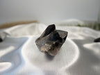 Treasured Energies raw smoky quartz crystal cluster with deep grey and brown tones on a white satin background.