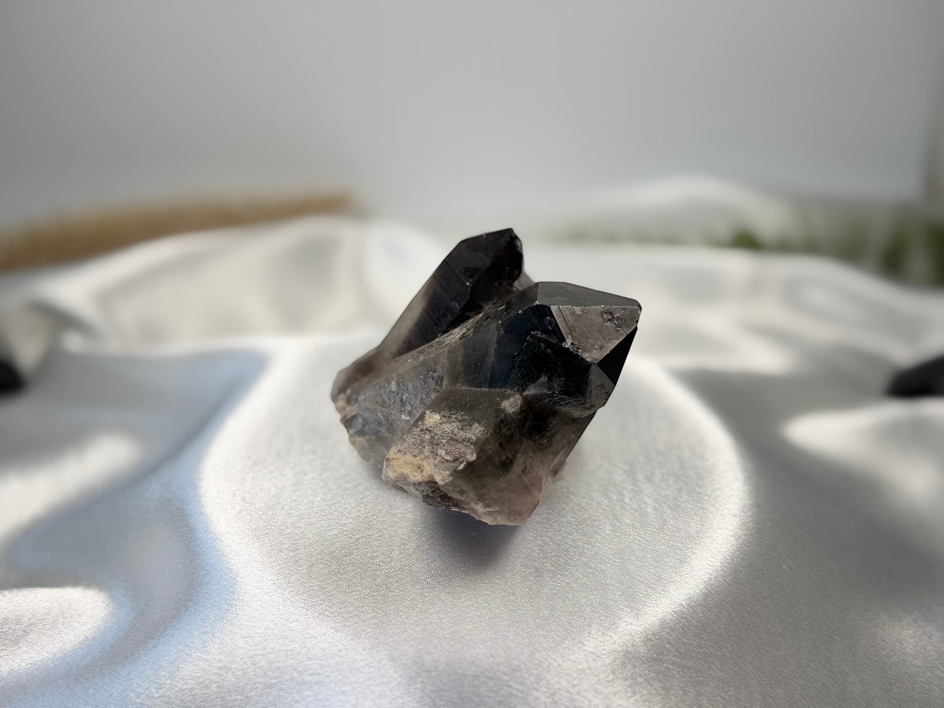 Treasured Energies raw smoky quartz crystal cluster with deep grey and brown tones on a white satin background.